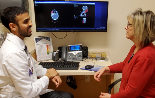 A female proton patient with blonde hair and an orange sweater sitting and smiling at a male proton therapy physician who is wearing a white lab coat and has brown hair and brown facial hair.