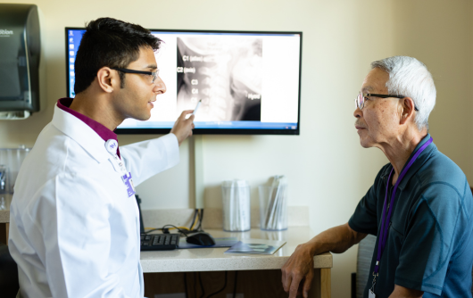 Proton Center doctor Akthar wearing a white lab coat and pointing to a computer screen of a spine while talking to an elderly Asian man.