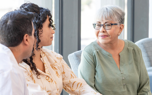 An elderly woman with grey hair, glasses and a green shirt talking with a young couple.