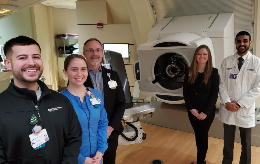 Five staff members from the Northwestern Medicine Proton Center standing around a Proton Therapy machine and smiling.
