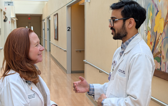 A male physician standing in a hallway wearing a white lab coat talking to a female staff member with red hair.