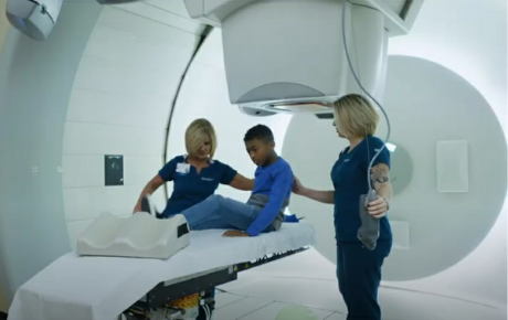 A pediatric proton therapy patient is helped onto the scan bed by two nurses.